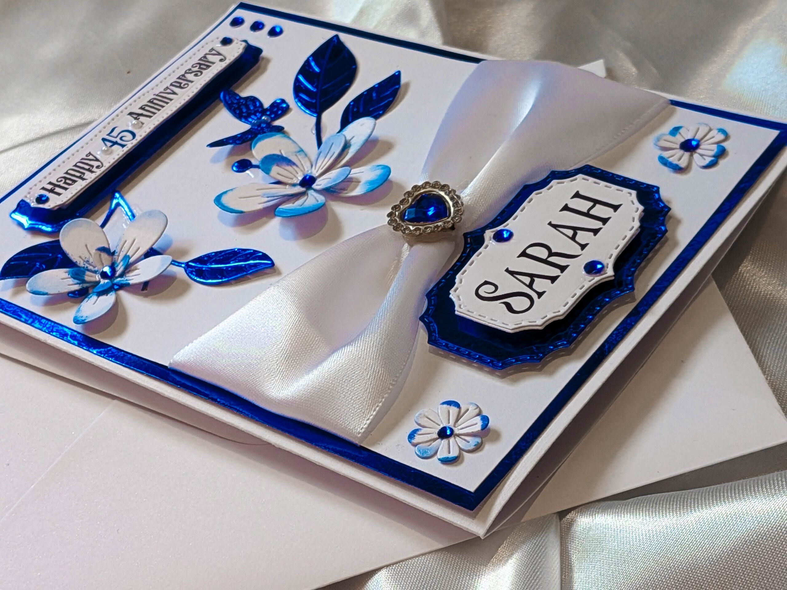 A full, three-quarter view of a personalised sapphire blue handmade card on a reflective satin background.
