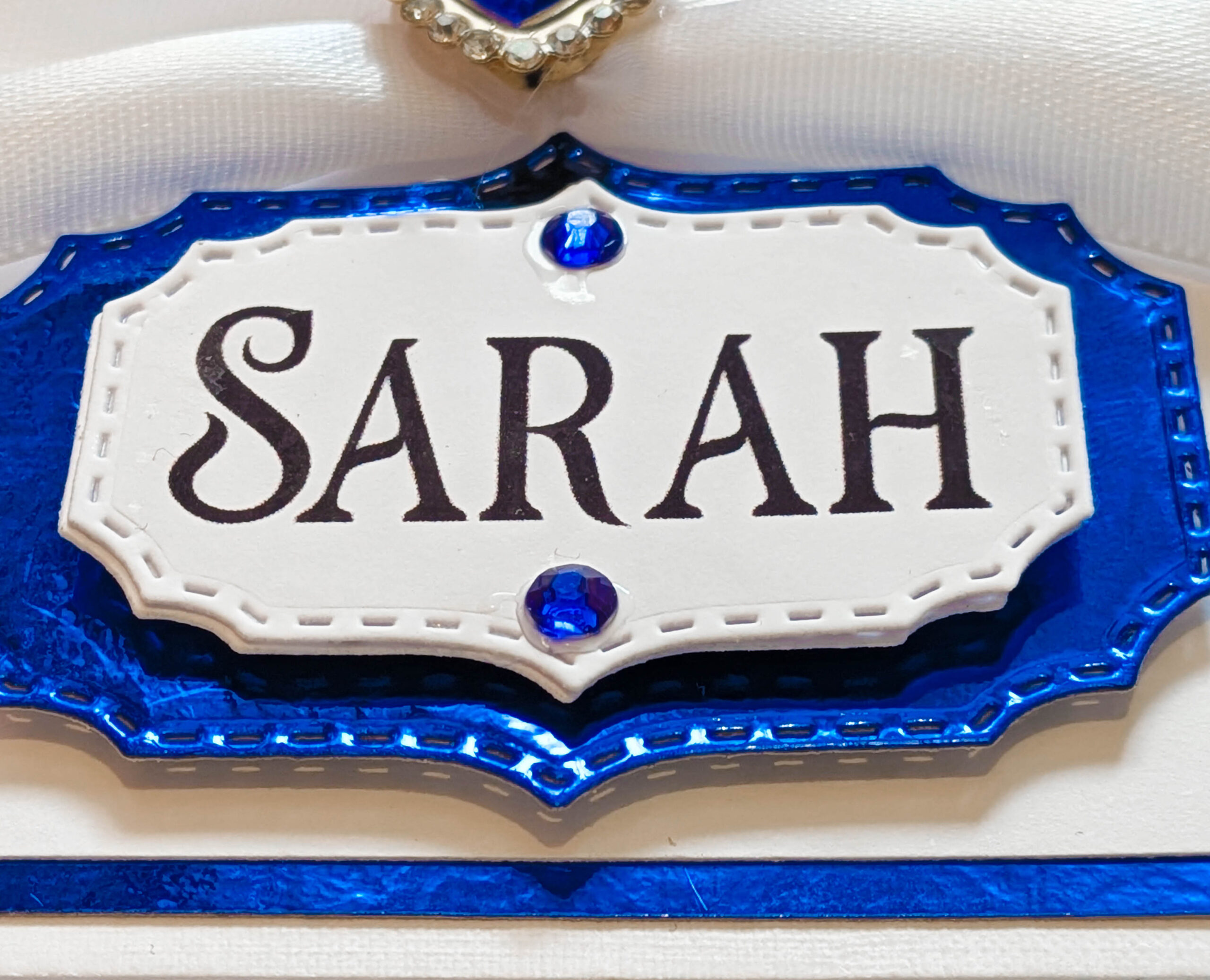 Close-up of a handmade 3D card showing a personalised name plaque with 'SARAH' below a white satin bow.