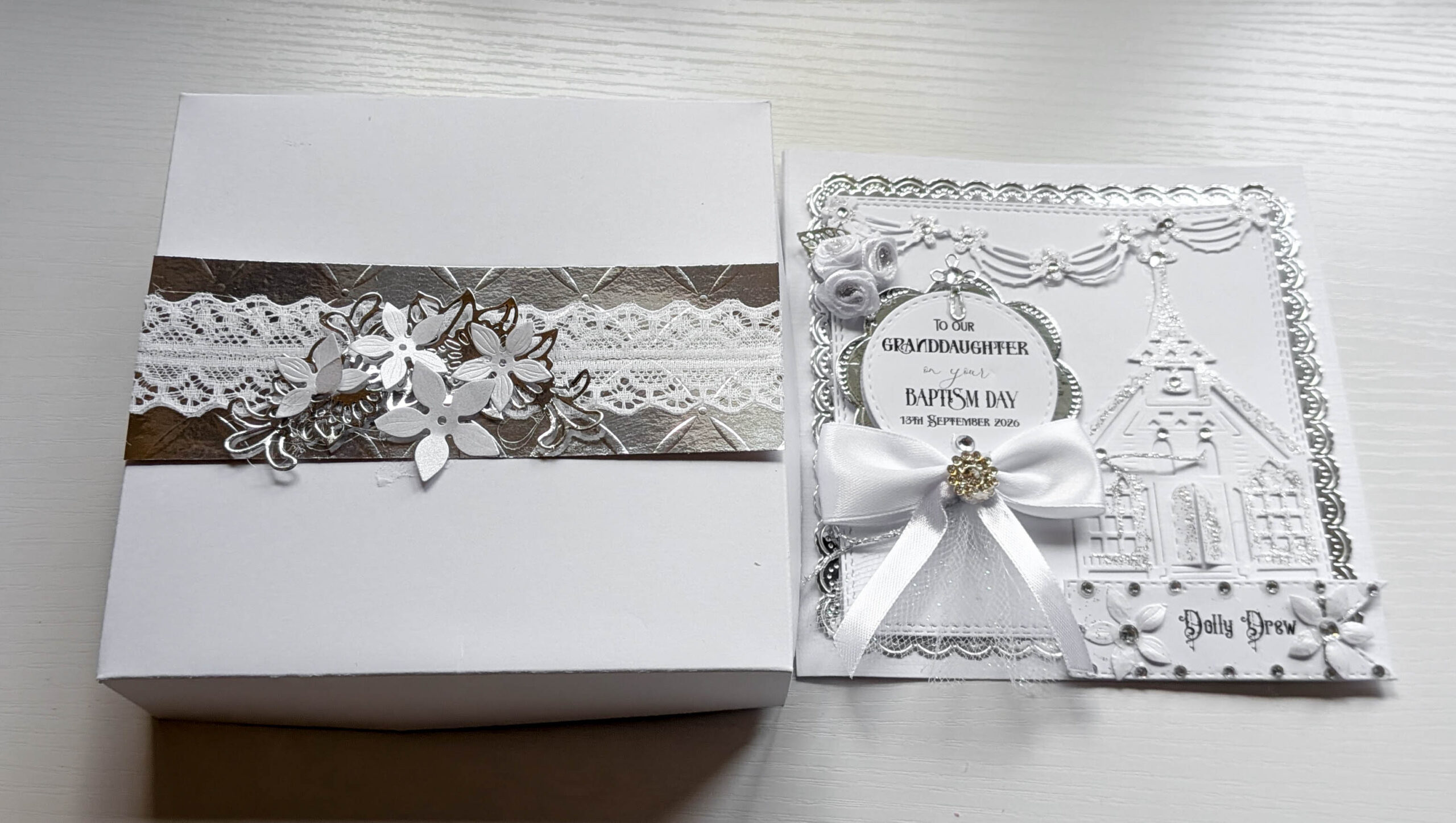 Handmade white christening card displayed partially inside its matching silver lace gift presentation box.